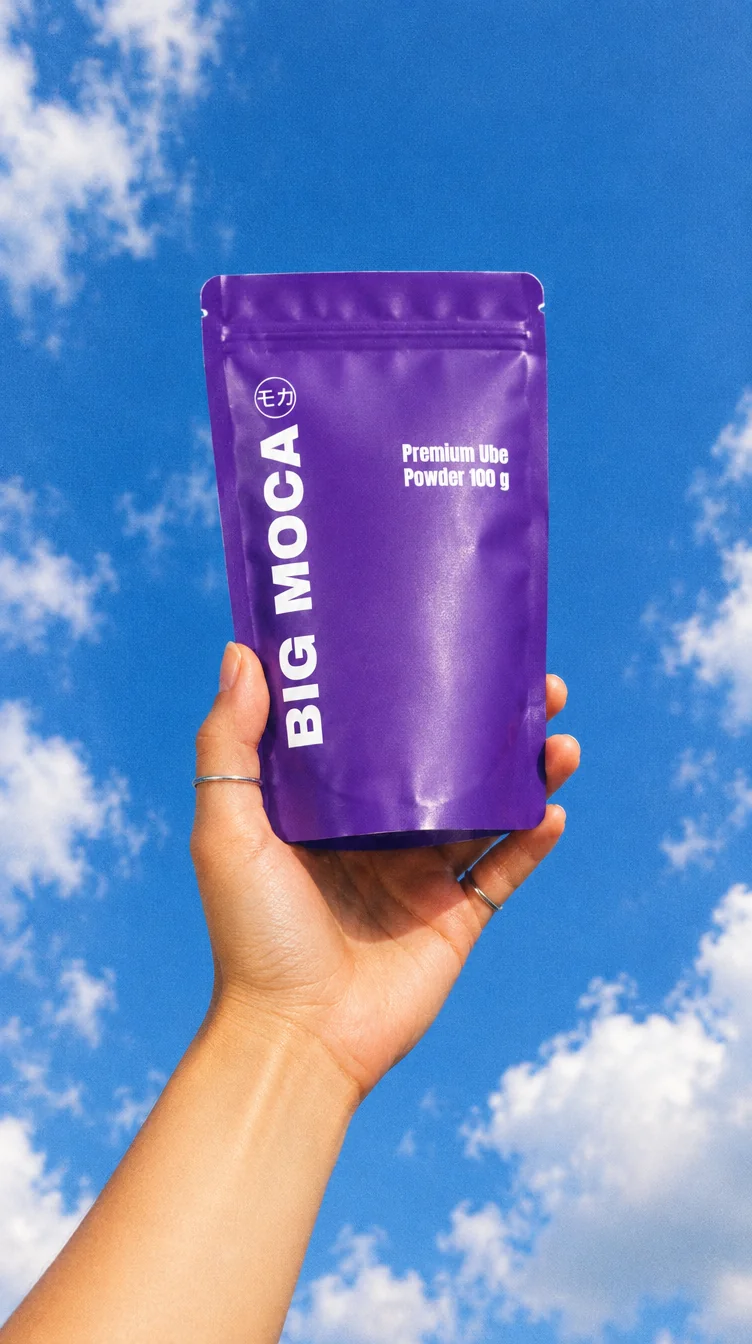 Hand houdt een zak Big Moca Premium Ube Powder omhoog tegen een blauwe lucht